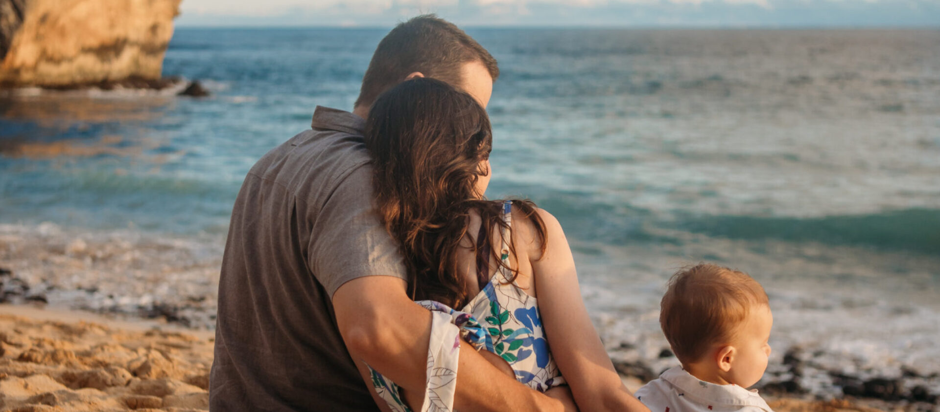 two adults and a baby site on a beach looking out at the ocean